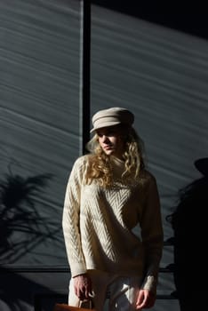 Studio portrait of beautiful woman with a curly blond hair, posing on gray background. Model wearing stylish cap, sweater and classic trousers