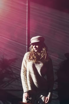 Studio portrait of beautiful woman with a curly blond hair, posing on gray background. Model wearing stylish cap, sweater and classic trousers