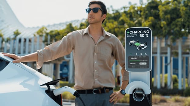 Young man put EV charger to recharge electric car's battery from charging station in city commercial parking lot. Rechargeable EV car for sustainable environmental friendly urban travel. Expedient