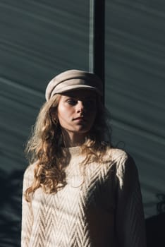 Studio portrait of beautiful woman with a curly blond hair, posing on gray background. Model wearing stylish cap, sweater and classic trousers