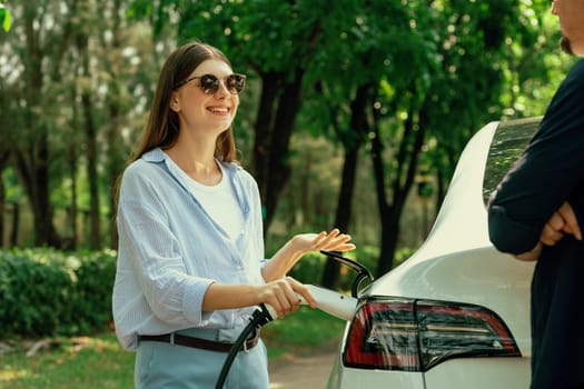 Lovely young couple wearing sun glasses recharging battery for electric car during road trip travel EV car in natural forest or national park. Eco friendly travel during vacation and holiday. Exalt