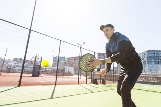 A padel player jump to the ball, good looking for posts and poster. Man with black racket playing a match in the open behind the net court outdoors. Professional sport concept with space for text. High quality photo