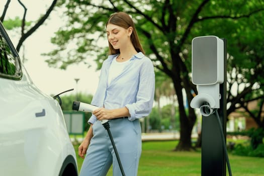 Young woman recharge EV electric vehicle's battery from EV charging station in outdoor green city park scenic. Eco friendly urban transport and commute with eco friendly EV car travel. Exalt