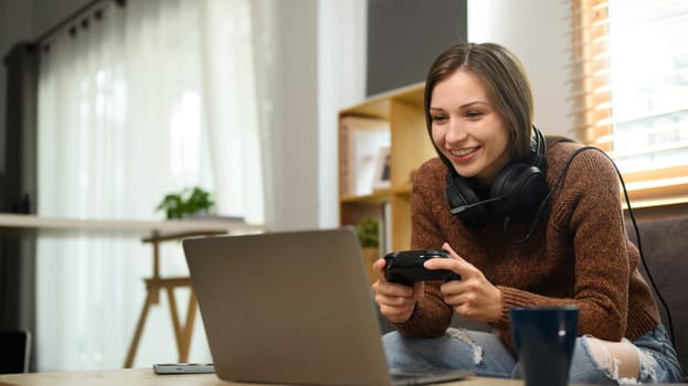 Joyful young caucasian woman in headset playing video games on laptop with joystick.