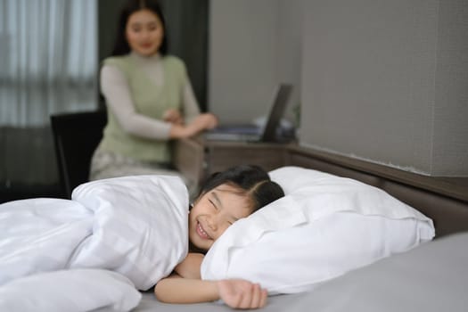 Playful child girl pretending to sleep on the bed with her mother sitting on background.