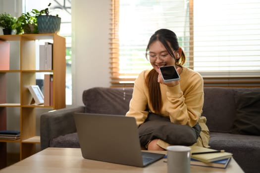 Shot of smiling young woman having phone conversation and using laptop on sofa.