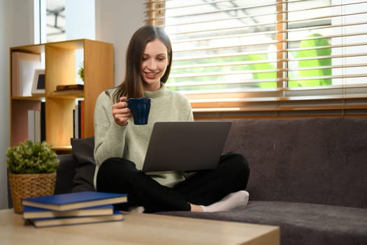 Beautiful caucasian lady drinking hot tea and using laptop in living room at home.