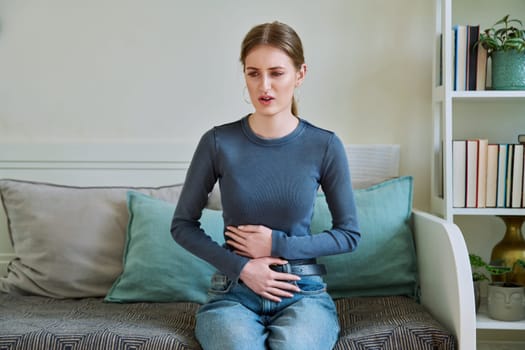 Young teenage female experiencing abdominal pain sitting at home. Stomach, menstrual cramps, gastrointestinal tract diseases, inflammatory processes in abdominal organs, kidneys, pelvis, youth, health