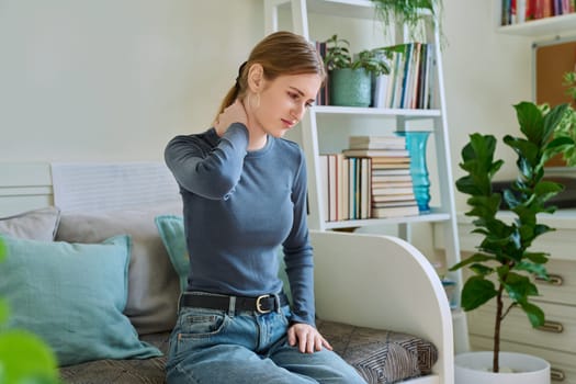 Young teenage female experiencing neck pain sitting on couch at home. Symptoms of the musculoskeletal system, neurological pathologies, acute infectious diseases