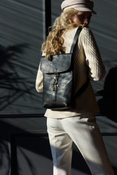 Studio portrait of beautiful woman with a curly blond hair holding brown bag, posing on gray background. Model wearing stylish cap, sweater and classic trousers