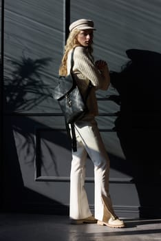 Studio portrait of beautiful woman with a curly blond hair holding brown bag, posing on gray background. Model wearing stylish cap, sweater and classic trousers