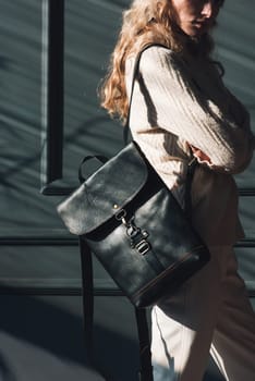 Studio portrait of beautiful woman with a curly blond hair holding brown bag, posing on gray background. Model wearing stylish cap, sweater and classic trousers