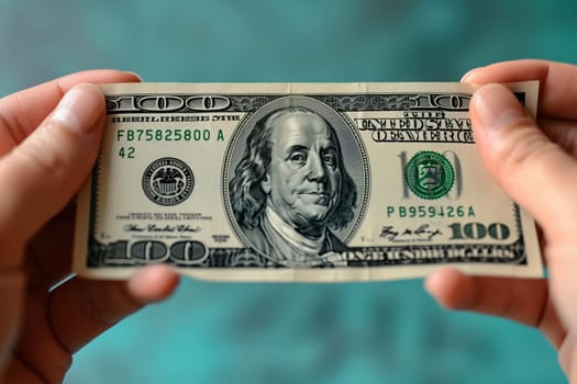 The persons hand is holding a crisp green 100 banknote. Their thumb and fingers are carefully handling the currency with a line of cash visible