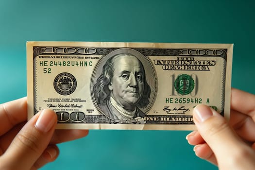 The person is holding a 100 dollar banknote in their hand, carefully handling the money with their fingers and thumb. The signature on the cash is clearly visible