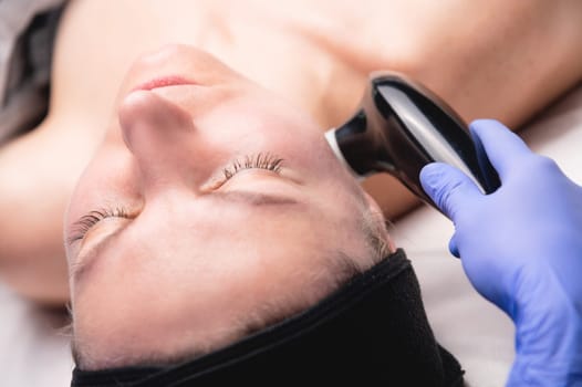 A middle-aged woman undergoes electrotherapeutic facial rejuvenation procedures in a cosmetologist's office