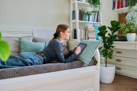 Young female teenager with digital tablet, stylus drawing illustrating lying on couch at home. Technologies, leisure, creativity, education, freelance work, lifestyle, youth concept