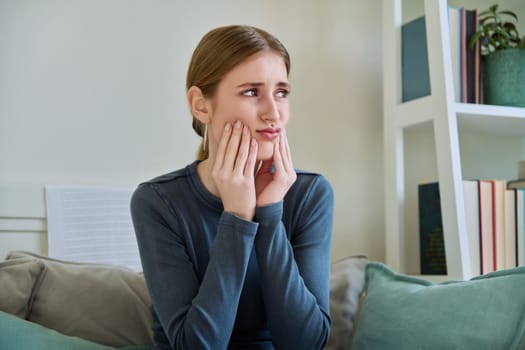 Young teenage female having toothache, suffering sad holding hands on face, sitting on couch at home. Teeth dentistry, dental, health, youth concept