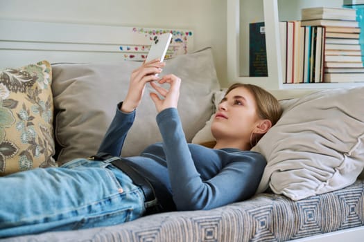 Happy smiling relaxed young female teenager lying on couch at home using smartphone for leisure communication. Youth, lifestyle, digital online technology, gadgets, internet