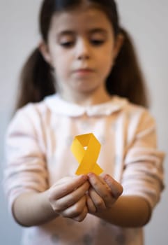 Beautiful little caucasian brunette girl with a sad face is holding out a yellow paper tape in her hands on a white background,close-up side view with depth of field. World childhood cancer day concept.