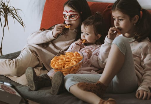 Three beautiful caucasian girls sisters are sitting on the sofa,eating corn rings and emotionally watching a movie on the tablet in the room in the evening,close-up side view.The concept of family watching movies.