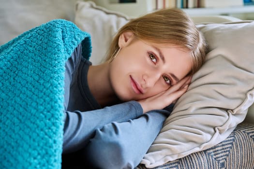 Relaxed resting young teenage female, looking at camera, lying on pillow under blanket, at home on couch. Health, beauty, rest calm peace, lifestyle, youth concept