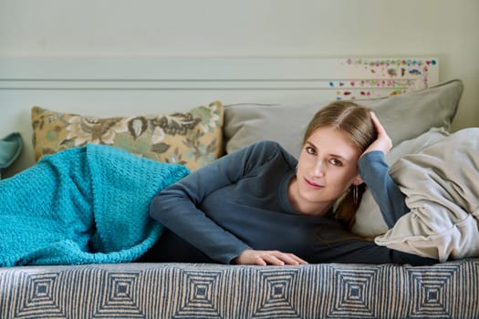 Beautiful smiling calm young teenage female relaxing lying under blanket at home on couch, looking at camera.