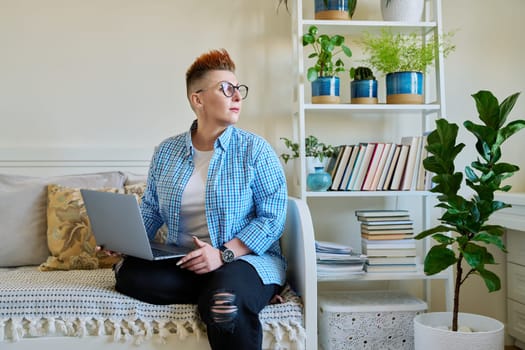 Middle aged woman sitting on couch at home using laptop. Serious female with red haircut in casual clothes working on computer, relaxing, communicating