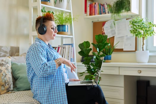 Middle aged woman freelancer in headphones using laptop computer sitting on couch at home. 40s female looking at screen video chat conference call online meeting training mentoring technology for work