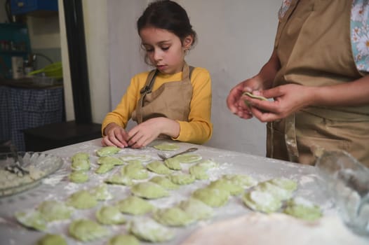Portrait Caucasian little child girl helping her mom in the kitchen, stuffing and molding dumplings, standing at floured table. Mom and daughter cooking together varennyky for family dinner at home.