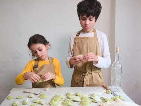 Authentic portrait of a teenage boy and school girl cooking together at home kitchen, making dumpling, helping their mom to prepare a dinner, standing at floured table, dressed in beige chef's apron