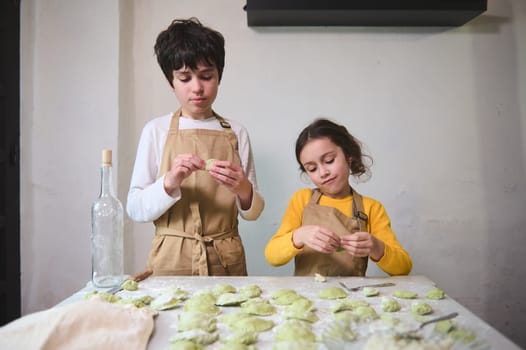 Two cute kids making dumplings in the home kitchen, dressed in beige chef's aprons. People. Culinary. Child learning cooking. Childhood. Domestic life
