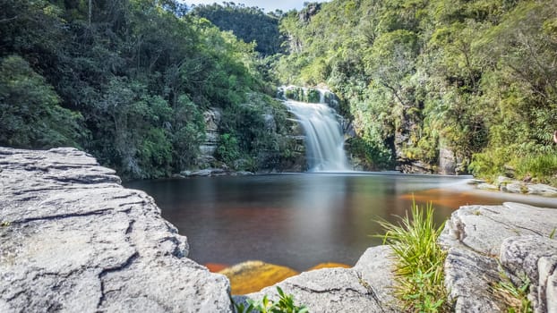 Tranquil waterfall flows into peaceful pond amidst lush greenery and stones.