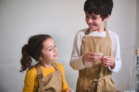 Authentic portrait of two adorable smiling kids making homemade dumplings at home kitchen, standing against white wall background