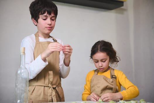 Cute kids in beige chef apron, making dumplings during a cooking class indoors, standing against a white wall background