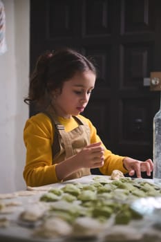 Authentic portrait of a serious little girl standing at kitchen table with molded raw ravioli and varennyki, molding dumplings for family dinner. People. Children. Food and cooking class concept