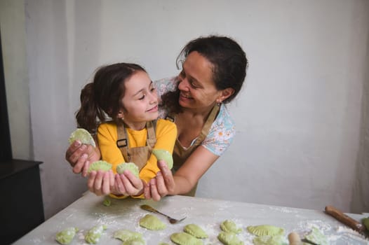 Smiling loving mother and her happy daughter looking at each other, holding sculpted homemade dumplings or Ukrainian varenyky, standing together at floured kitchen table, against white wall background