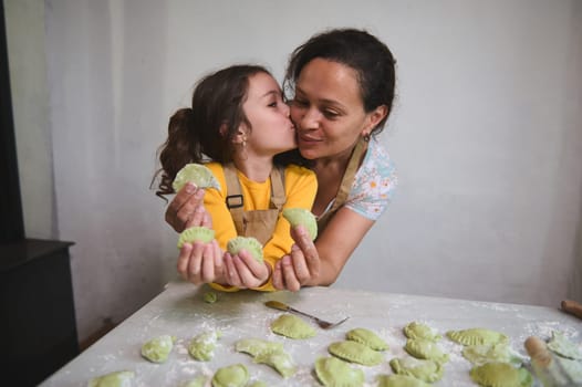 Mom and daughter have fun while cooking dumpling together. Kid girl kissing her mother, happy to learn the traditional Ukrainian culinary, showing molded varennyky to camera, standing at floured table