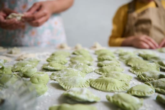 Selective focus on molded homemade dumplings on floured table, against the background of young mom and daughter cooking together. Ravioli. Pelmeni. Varennyky stuffed with mashed potatoes