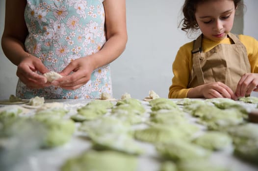 Selective focus on sculpted homemade dumplings on floured table, against the background of young mom and daughter cooking together