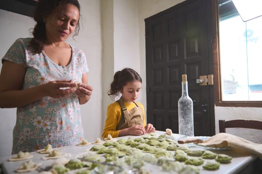Authentic beautiful young woman, mother and her little kid, daughter in the rural kitchen, sculpting dumplings from dough with mashed potatoes filling. Cooking delicious homemade vegetarian dumplings