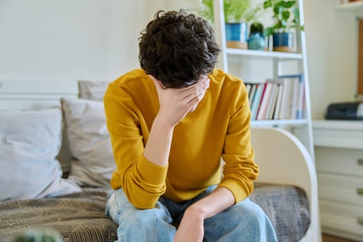 Sad upset tired young man sitting on couch at home, touching his head with hands. Health problems, headaches, troubles, difficulties in study, family relationships, mental health, stress, depression