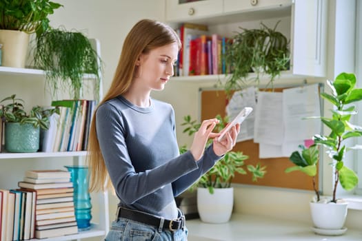 Happy smiling female teenager using smartphone, standing at home, girl 16,17, 18 years old texting reading messages. Modern digital technologies for communication, leisure, learning, shopping