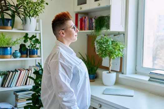 Profile portrait of serious middle-aged woman in glasses with red haircut looking at window in home interior, copy space. Mature people, lifestyle, health, life concept
