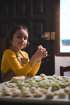 Caucasian beautiful child girl, little chef in beige apron, holding a homemade dumpling, smiles looking at camera, standing at floured table with molded ravioli, pelmeni, vareniki, hinkali
