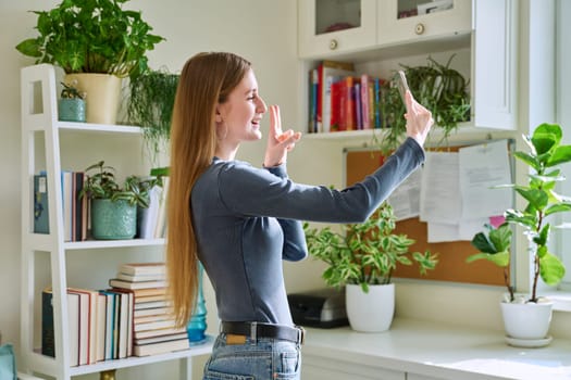 Fun grimacing happy young teenage female taking selfie photo on smartphone, in home interior. Joy, fun, youth, lifestyle concept