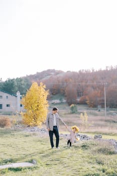 Dad looks at the little girl in a wreath of yellow leaves as he walks holding hands across the sunny lawn. High quality photo
