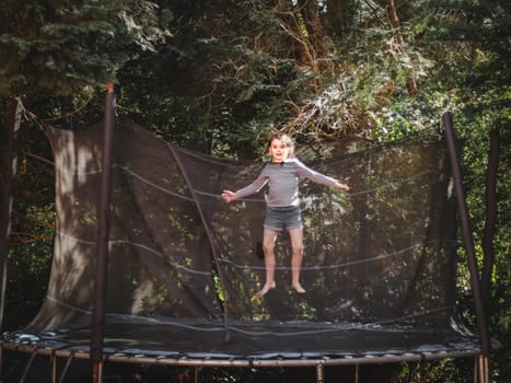 A beautiful caucasian girl in shorts and a sleeved T-shirt jumps on a trampoline, frozen in a straight position with outstretched arms in the garden of a house among trees, close-up side view. Concept at home, home sports, happy childhood, children entertainment.