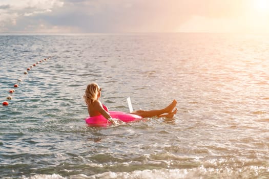 Woman works on laptop in sea. Freelancer, blond woman in sunglases floating on an inflatable big pink donut with a laptop in the sea at sunset. People summer vacation rest lifestyle concept