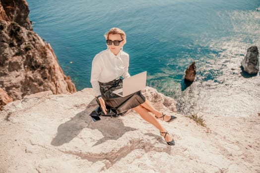 Business woman on nature in white shirt and black skirt. She works with an iPad in the open air with a beautiful view of the sea. The concept of remote work