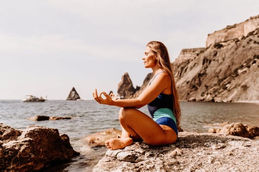 Yoga on the beach. A happy woman meditating in a yoga pose on the beach, surrounded by the ocean and rock mountains, promoting a healthy lifestyle outdoors in nature, and inspiring fitness concept